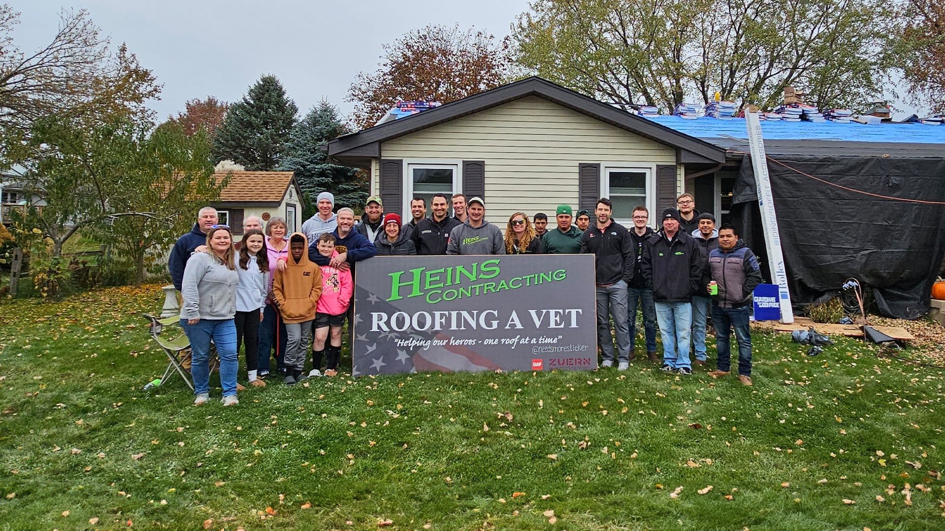 The Heins Contracting team and community members stand behind a "Roofing a Vet" banner.